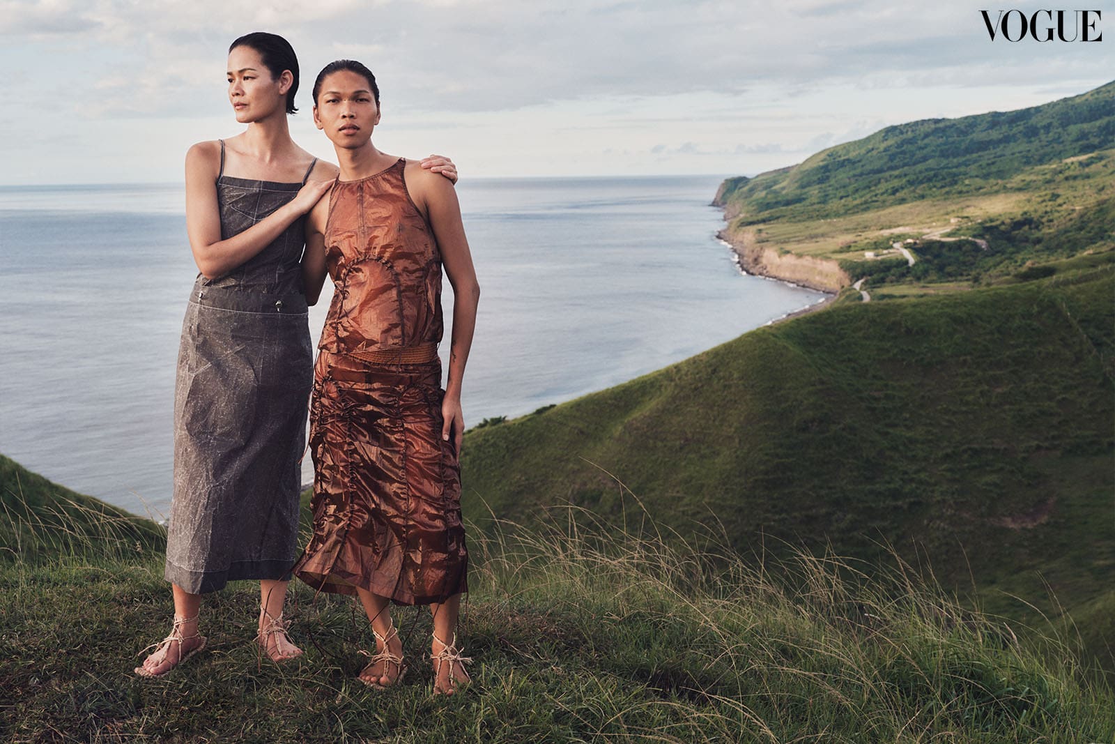 Jo Ann wears a coated gray vinyl apron dress while Lukresia wears a caramel-colored “tech organza” ensemble, both from ECKHAUS LATTA. Both wear Ivatan Tukap rope sandals.
