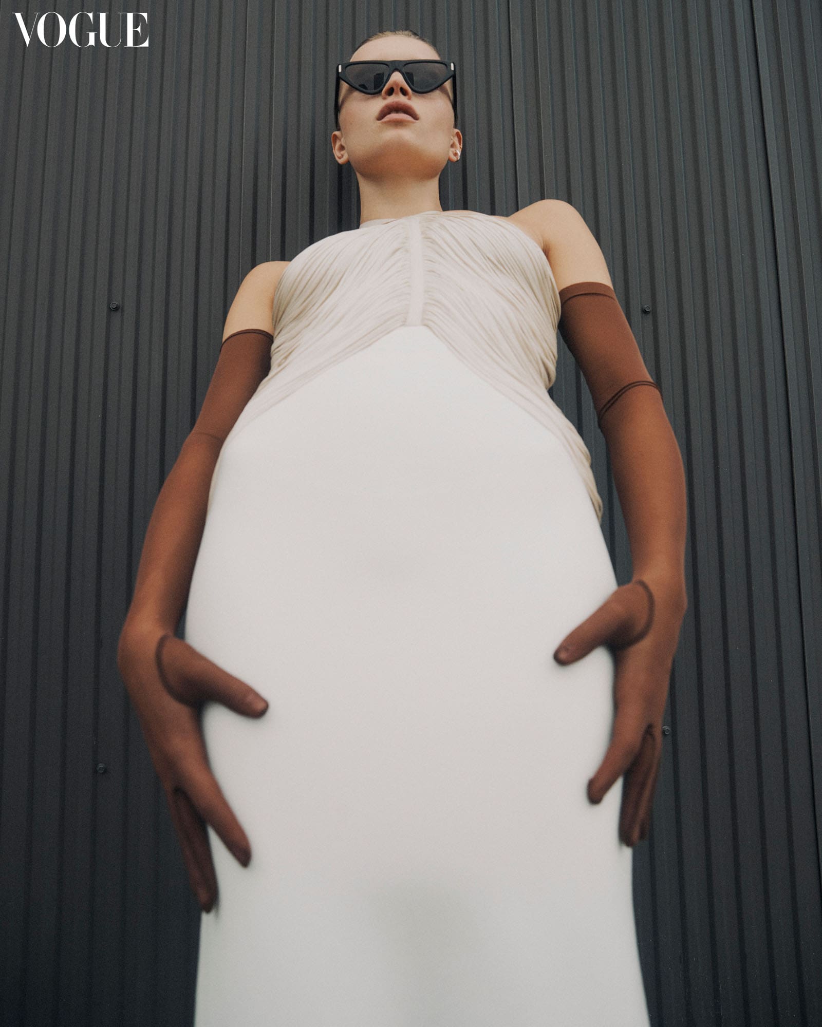 Model posing against a black textured wall wearing Michelle Ochs white dress, brown gloves, and black sunglasses