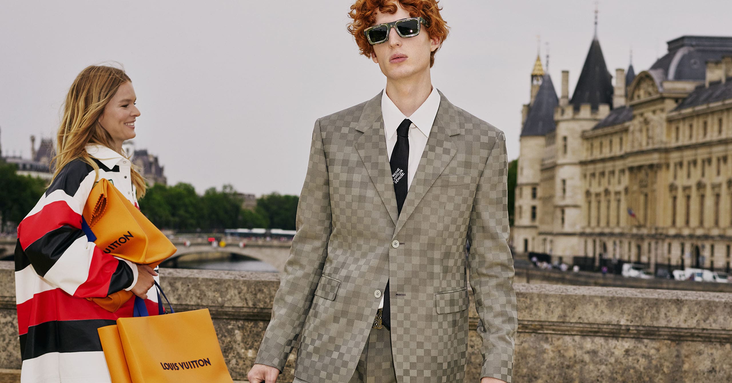 A Louis Vuitton modeling with a grey checkered suit with a trunk black Louis Vuitton bag