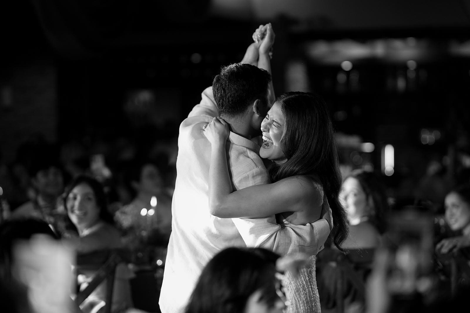 A black-and-white image captures a tender moment between Kiana Valenciano and Sandro Tolentino during their wedding celebration. The couple embraces while dancing, with Kiana laughing joyfully, her arm wrapped around Sandro. The scene is filled with emotion, illuminated softly in a room of smiling guests in the background.