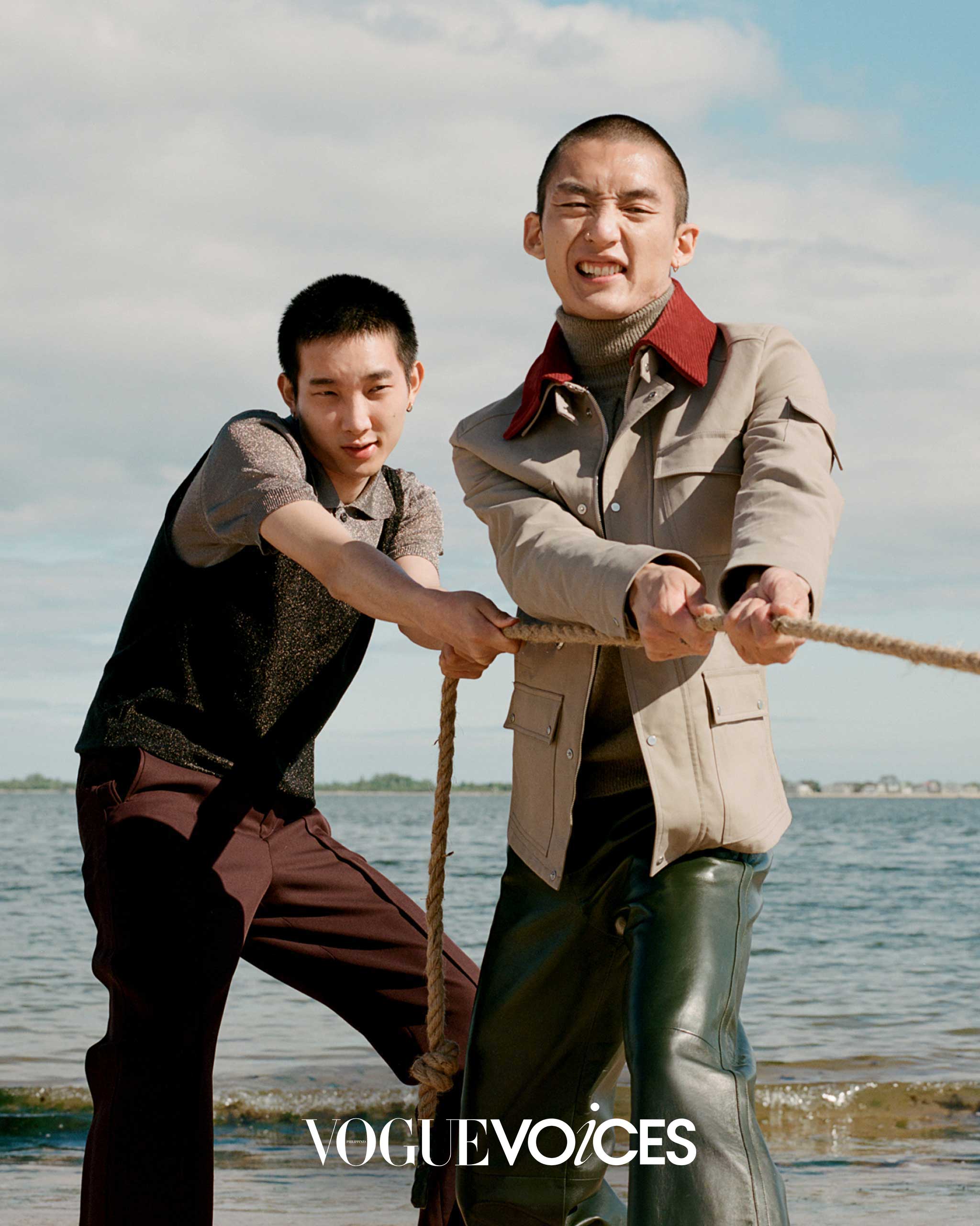 Two individuals are seen outdoors near the water, engaging in a tug-of-war. The person on the left wears a sleeveless sweater layered over a short-sleeve shirt and dark trousers, while the person on the right wears a beige jacket with a red collar, a turtleneck, and green leather pants.