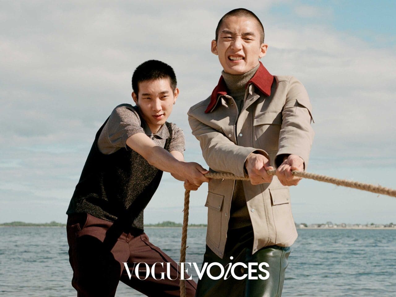 Two individuals are seen outdoors near the water, engaging in a tug-of-war. The person on the left wears a sleeveless sweater layered over a short-sleeve shirt and dark trousers, while the person on the right wears a beige jacket with a red collar, a turtleneck, and green leather pants.