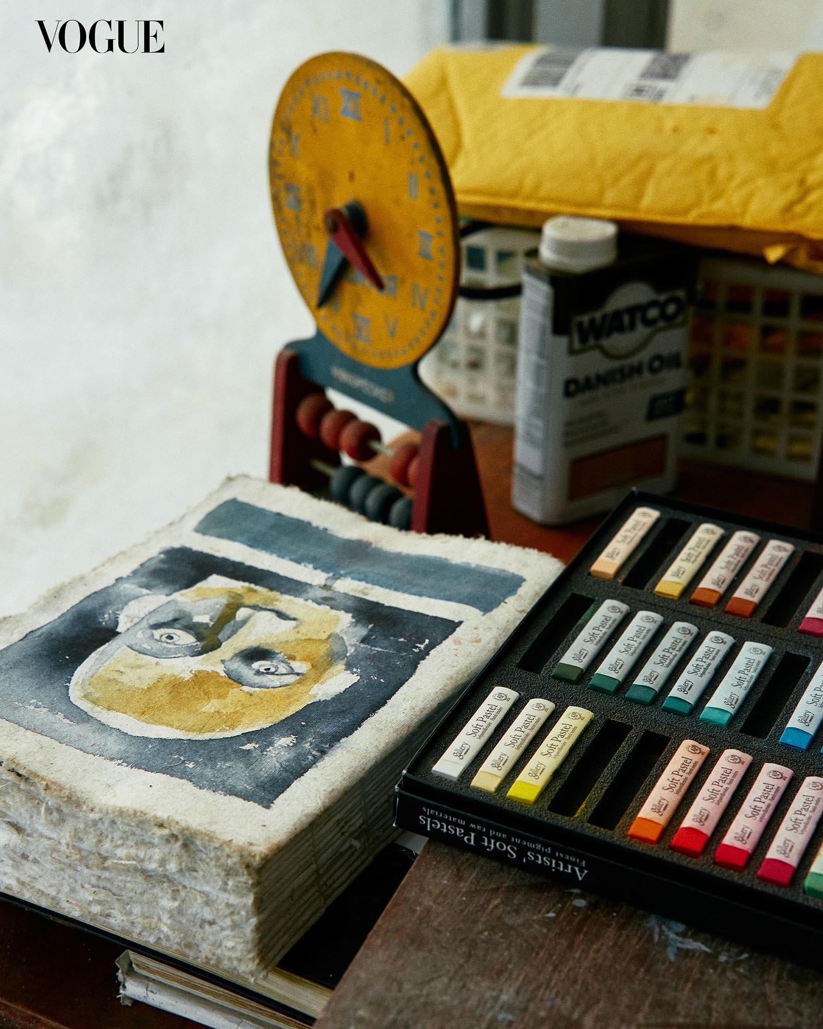 Close up of art materials, a set of soft pastels with a few empty containers, a sketchbook, and a bottle of paint thinner.