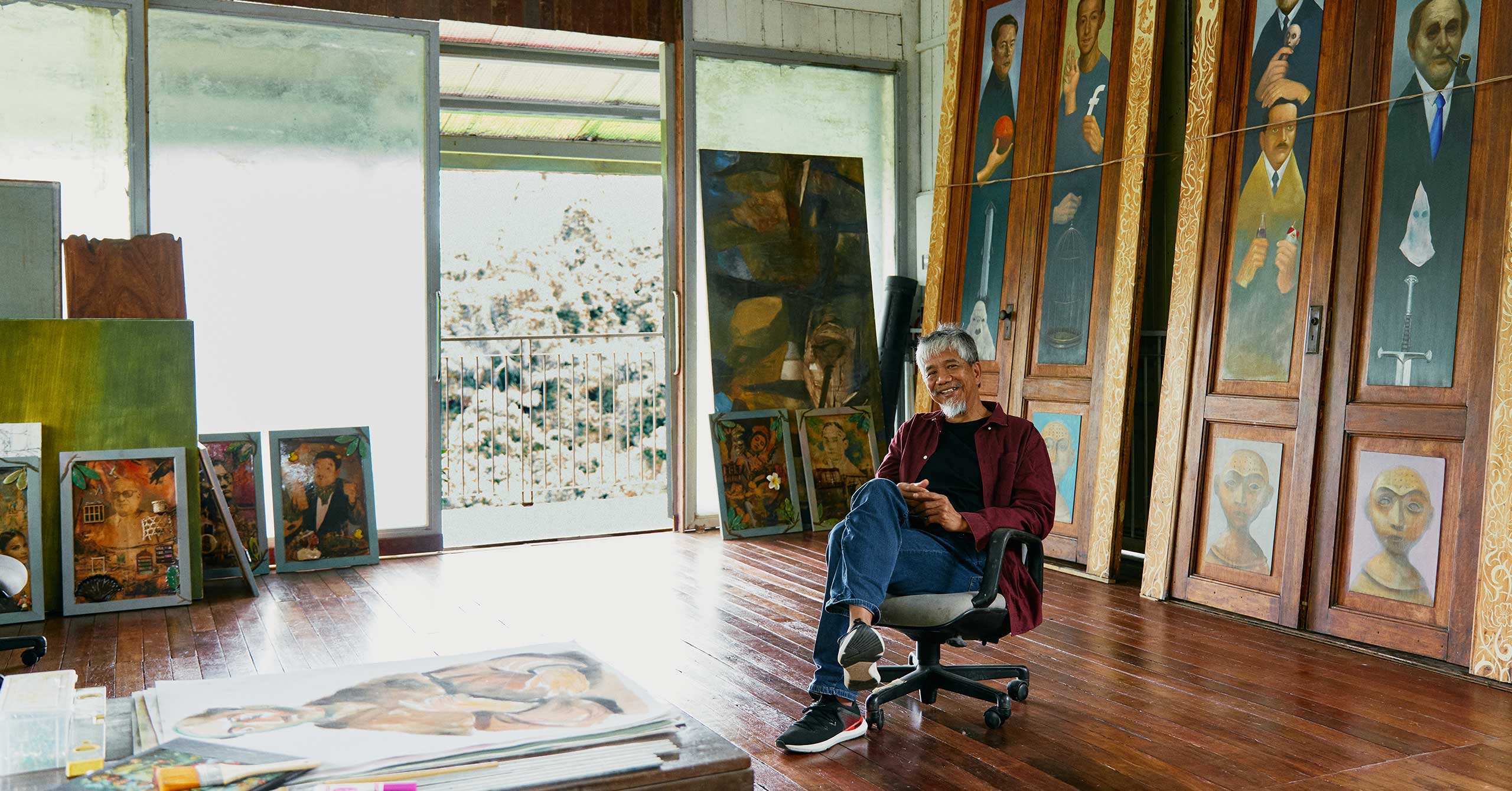 The artist Manny Garibay sits on a swivel office chair in front of his two paintings.