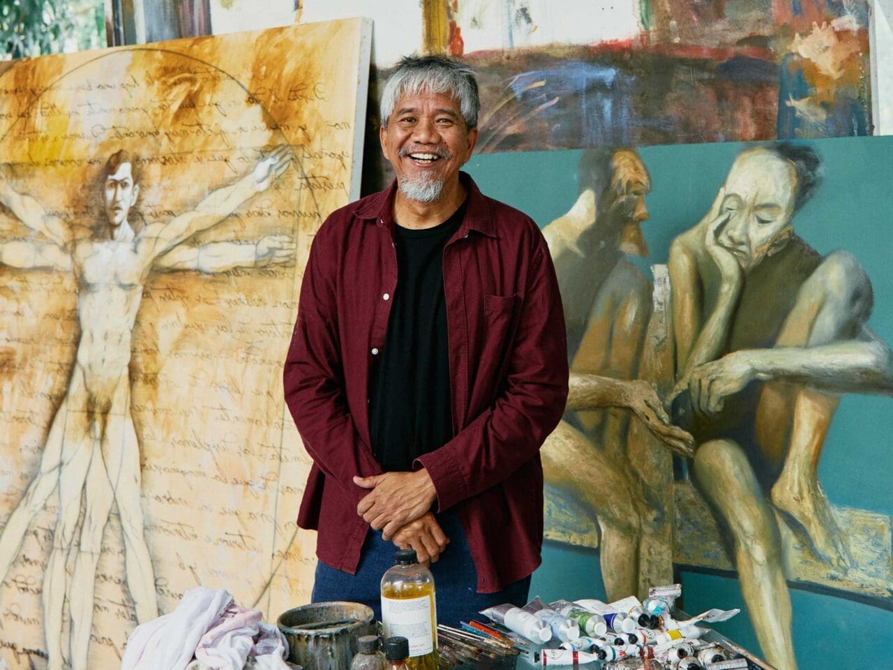 The artist Manny Garibay sits on a swivel office chair in front of his two paintings.